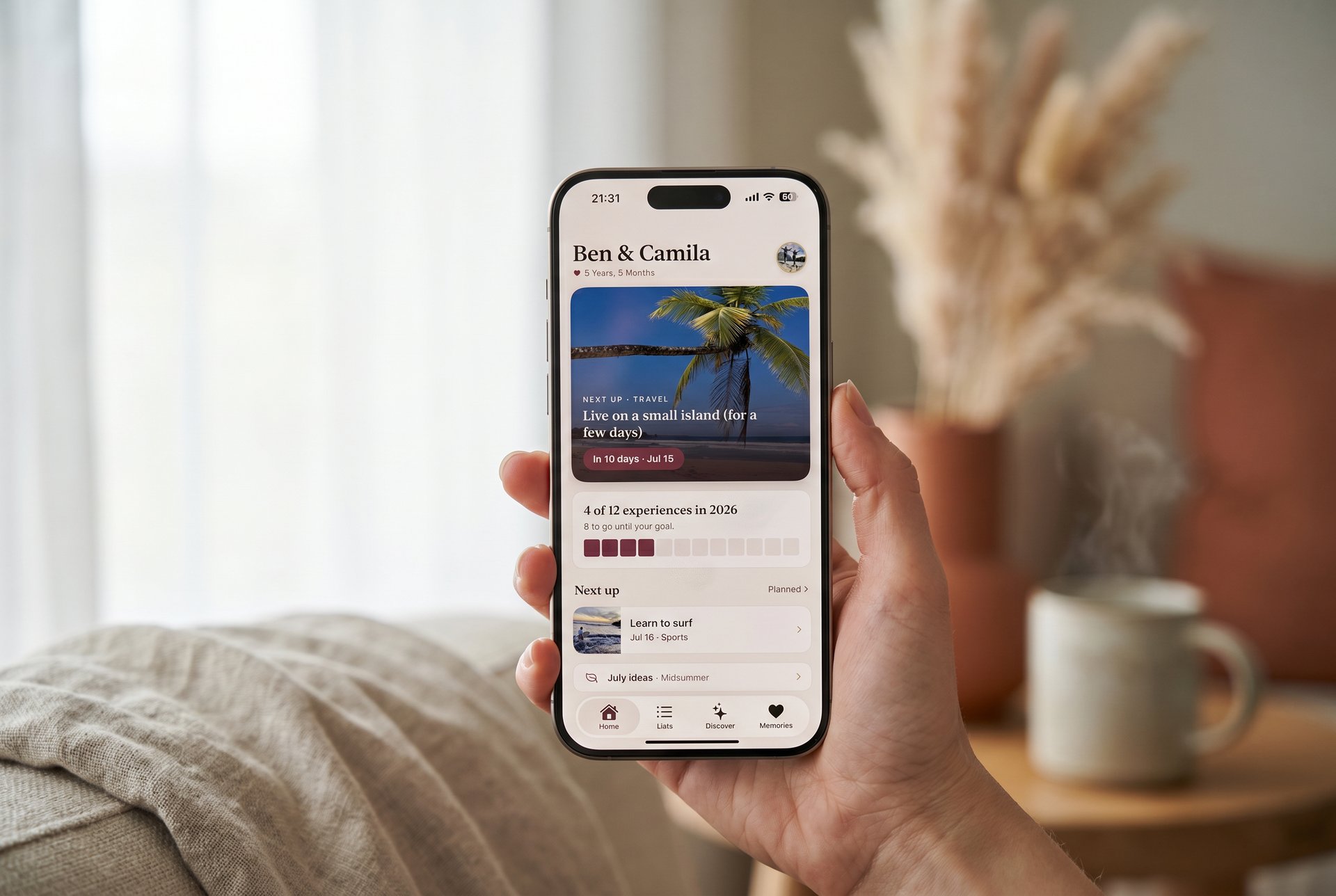 A hand holding an iPhone showing the Korby app in a warm, sunlit living room